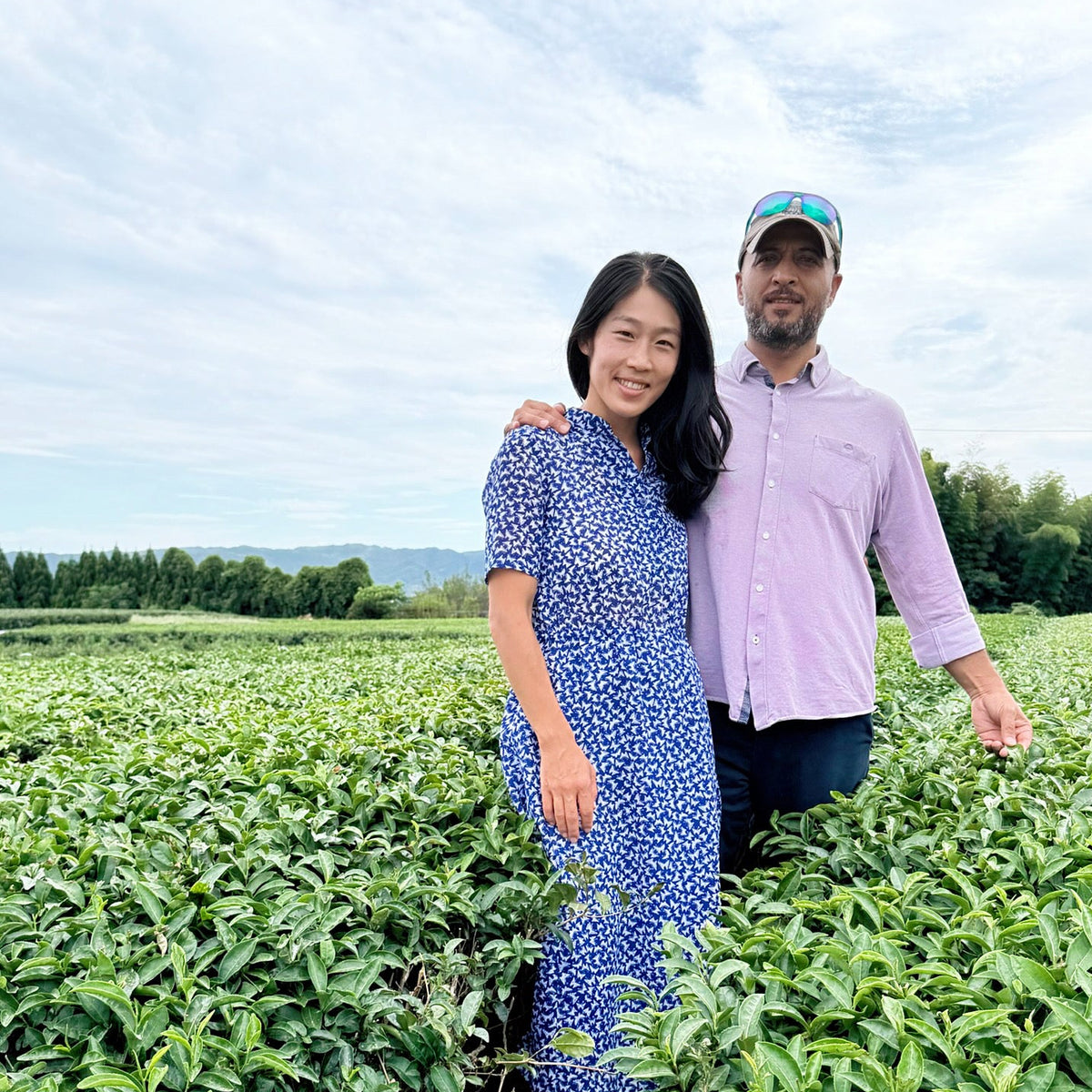 A man and a woman standing together in a lush green tea plantation under a cloudy sky. The woman wears a blue and white patterned dress, and the man wears a light purple button-down shirt and a baseball cap.